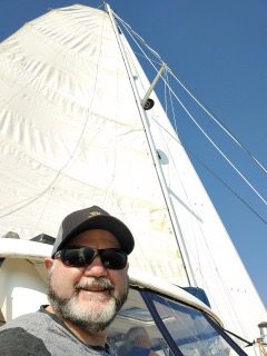 A man in sunglasses and hat standing on the front of a sail boat.